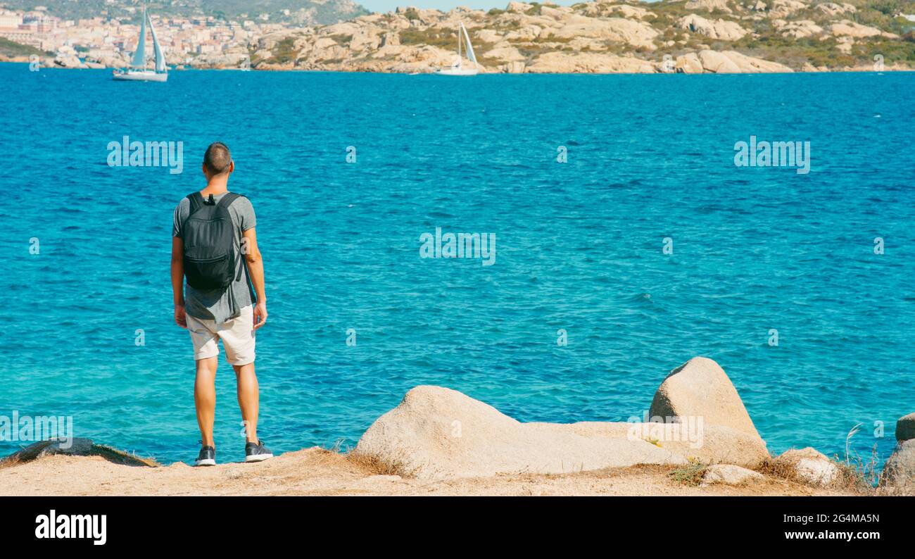 Il giovane, che porta con sé uno zaino, osserva la costa del Mediterraneo in Sardegna, Italia, guardando verso le isole la Maddalena e Santo Stefano Foto Stock