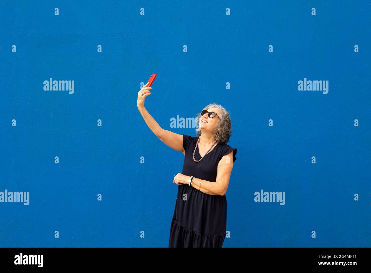 Donna anziana con capelli grigi che prende un selfie con uno smartphone rosso contro una parete blu Foto Stock