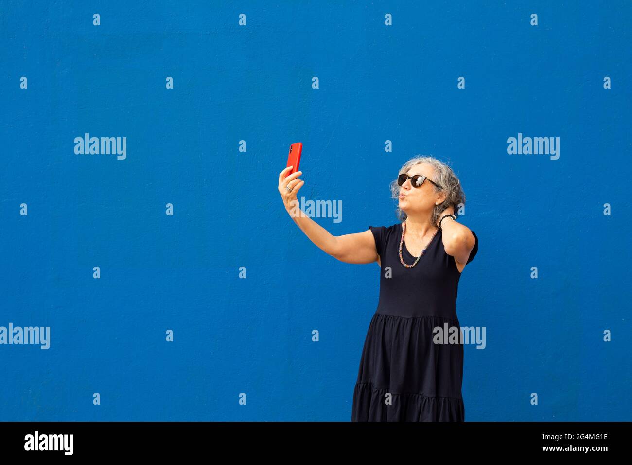 Donna anziana con capelli grigi che prende un selfie con uno smartphone rosso contro una parete blu Foto Stock