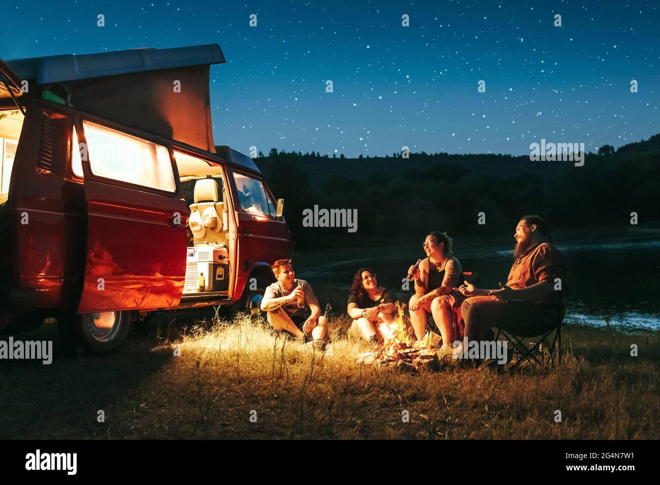 Allegro gruppo di amici che si divertono a campeggiare vicino al lago e pulmino durante la notte stellata Foto Stock