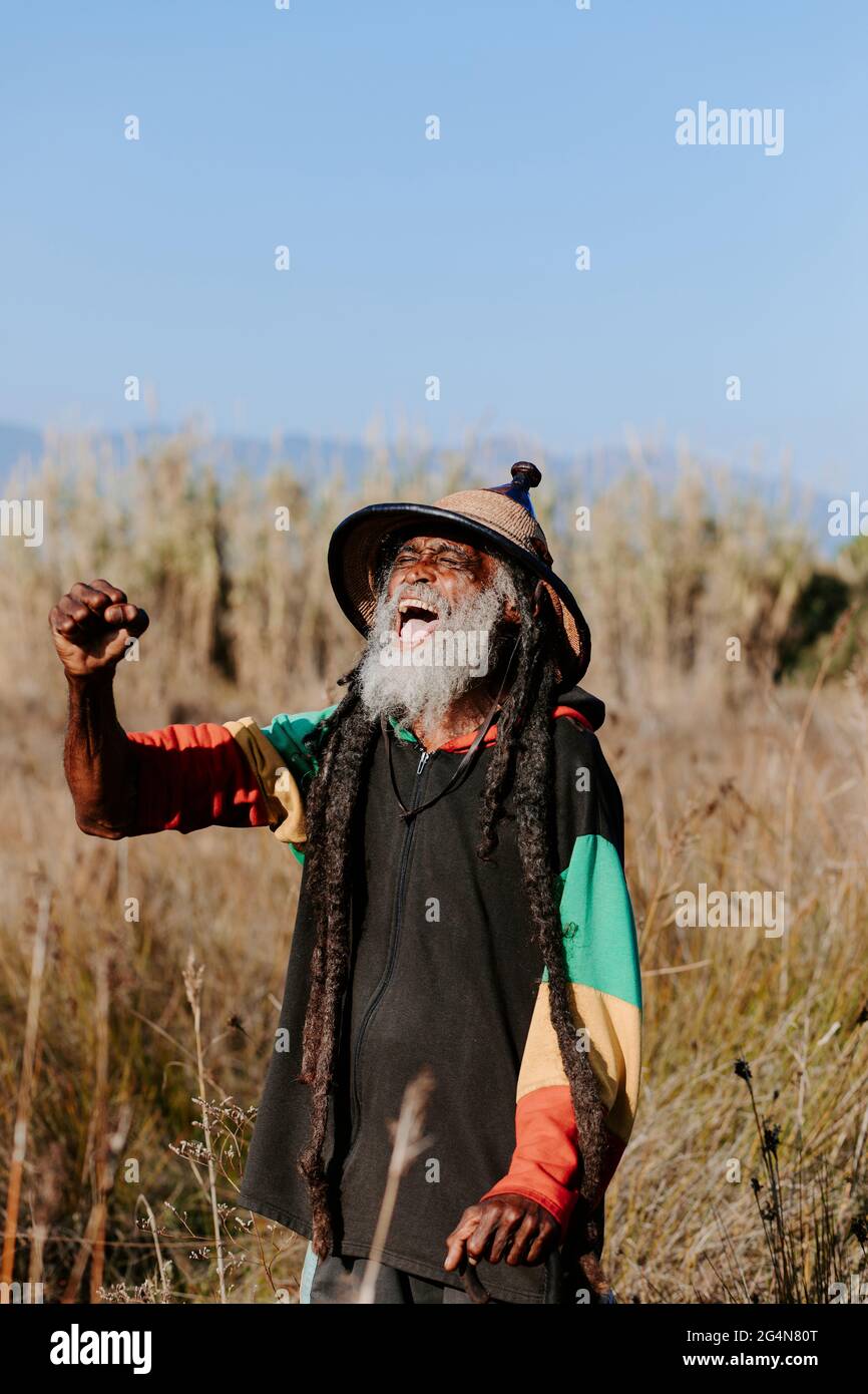 Allegro vecchio rastafari etnico con tempri con gli occhi e pugno ha chiuso celebrando la vittoria mentre si è in piedi in un prato asciutto nella natura Foto Stock