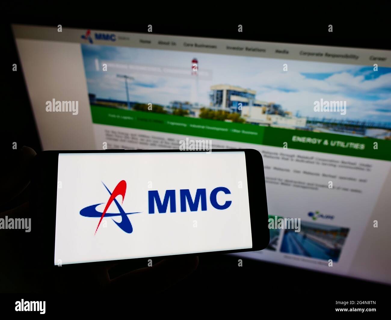 Persona che tiene il cellulare con il logo della società malese MMC Corporation Berhad sullo schermo di fronte alla pagina web aziendale. Mettere a fuoco il display del telefono. Foto Stock