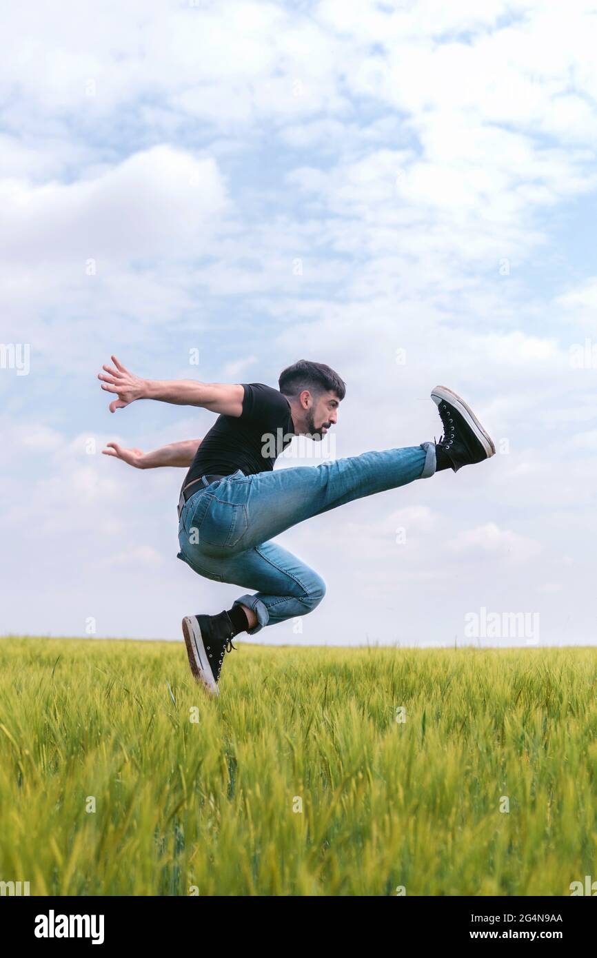Vista laterale uomo in denim salto con gamba rialzata sopra erba alta in un campo cupo Foto Stock