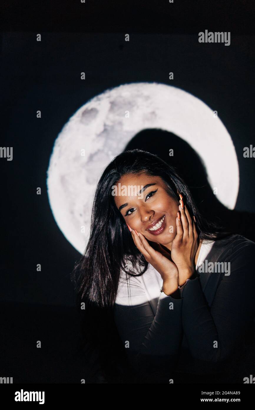 Giovane ragazza ispanica allegra che guarda le guance che toccano la fotocamera mentre riposa sotto la proiezione della luna di notte Foto Stock