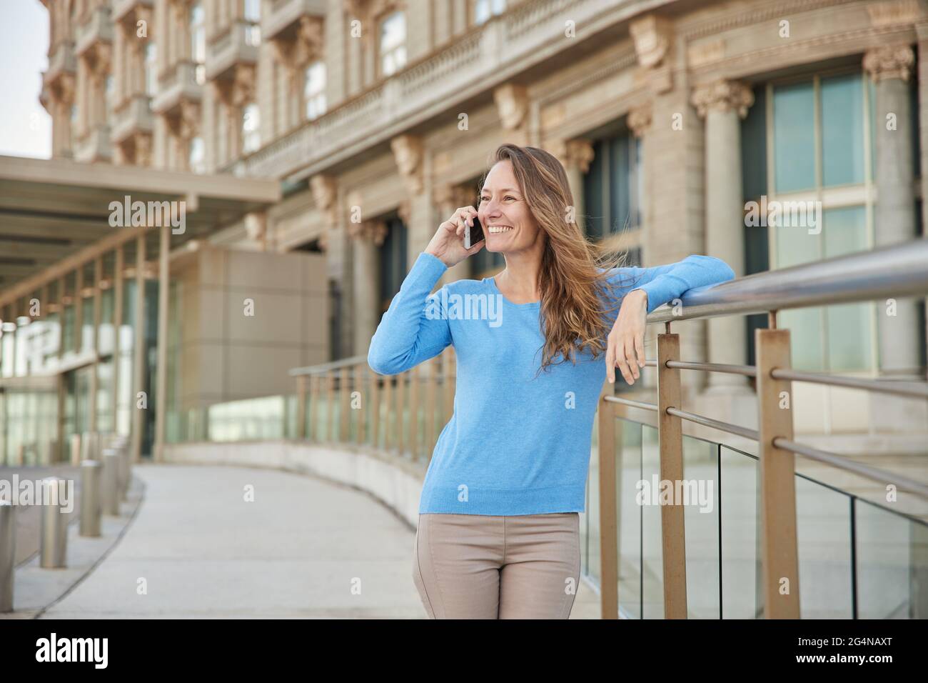 Donna adulta sorridente in abiti informali in piedi vicino recinzione e vecchio edificio mentre si parla telefono nel quartiere della città in luce del giorno Foto Stock