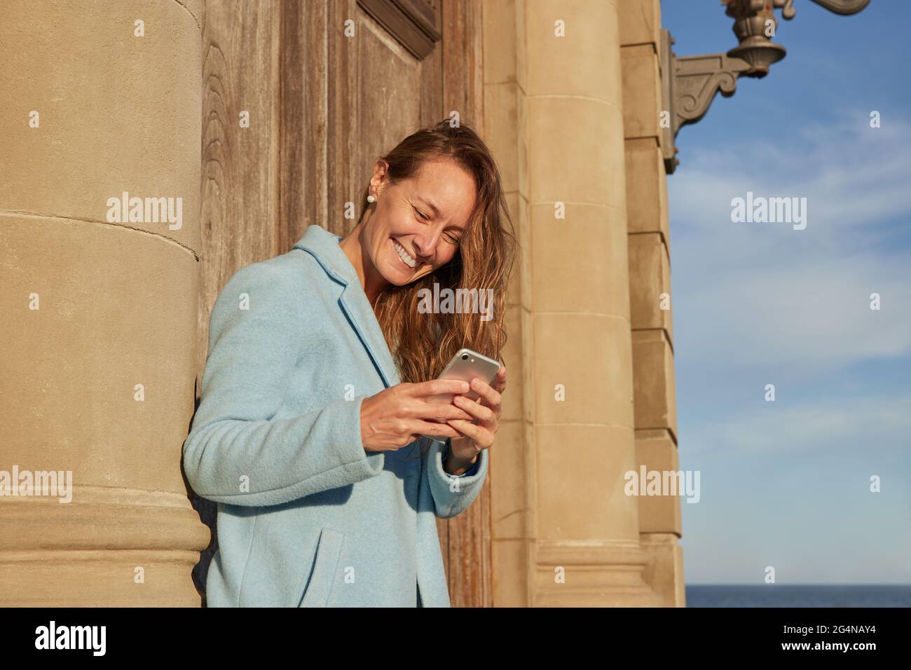 Felice adulto Donna in cappotto blu appoggiato su vecchio edificio mentre navigando sul cellulare nel quartiere della città in giorno di sole sotto cielo nuvoloso blu Foto Stock