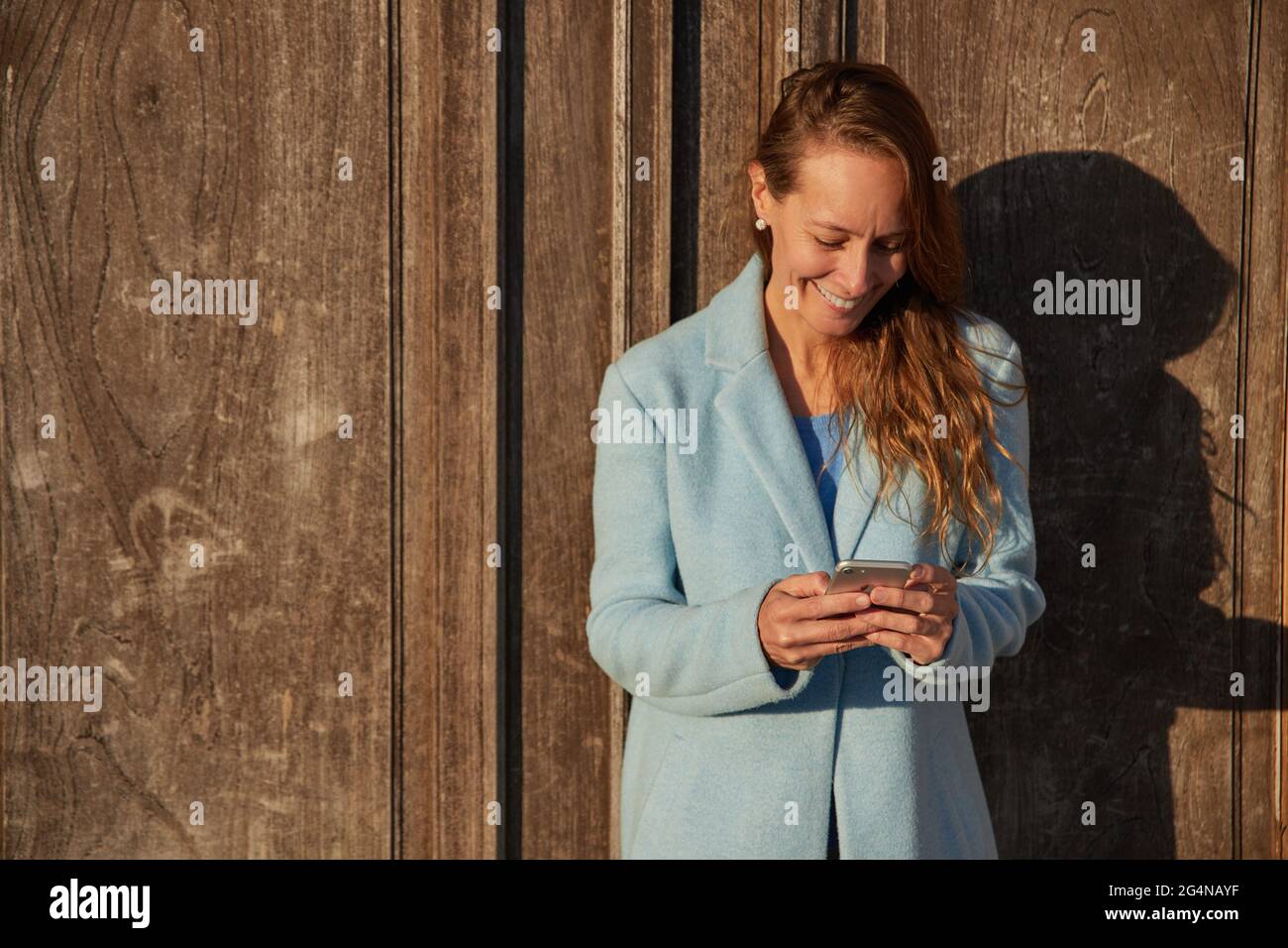 Felice adulto Donna in cappotto blu appoggiata su vecchio edificio mentre navigando sul cellulare nel quartiere della città in giorno di sole Foto Stock