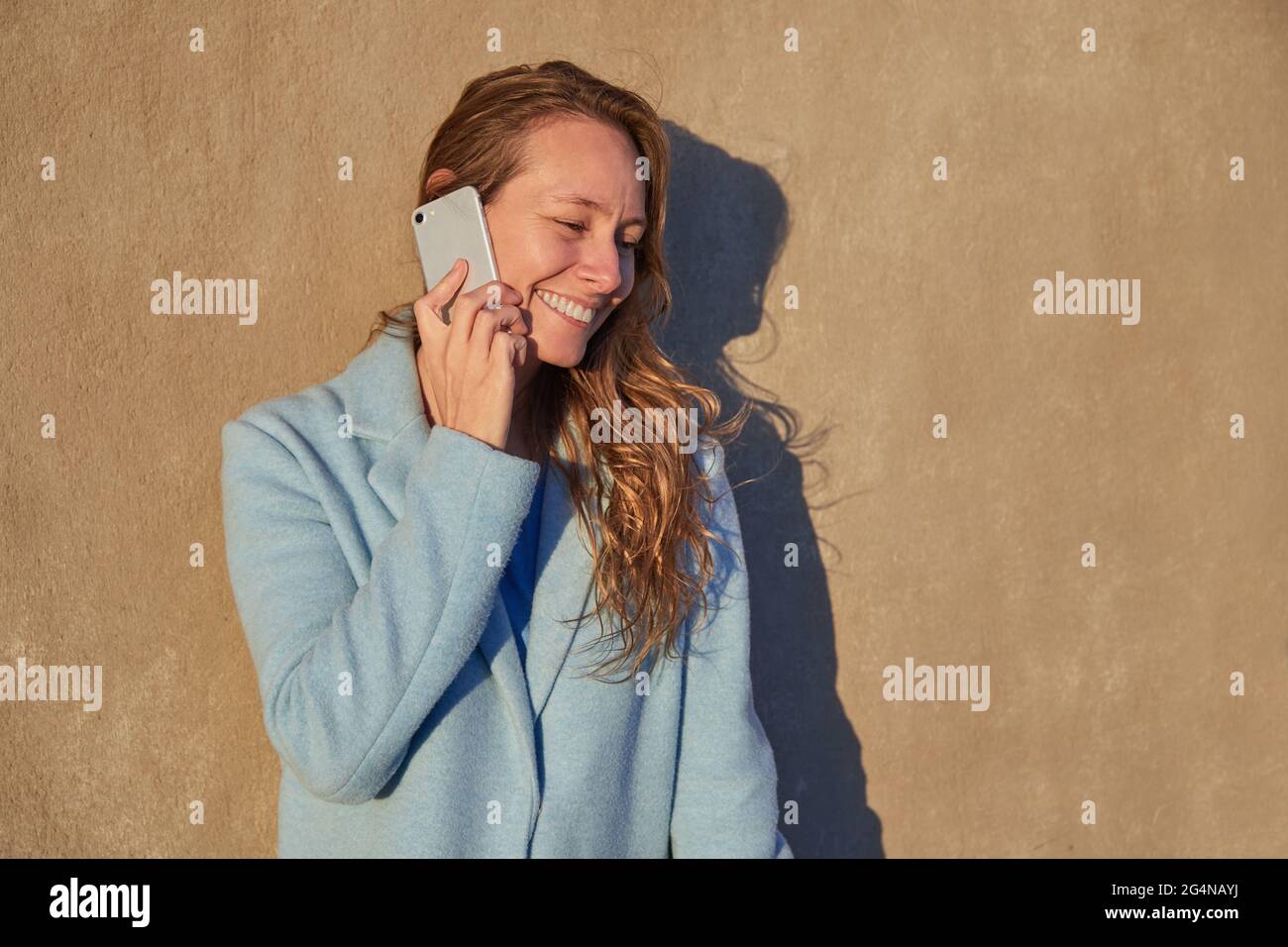 Felice adulto Donna in cappotto blu appoggiato sulla vecchia parete mentre parlando sul cellulare nel quartiere della città in giorno di sole Foto Stock
