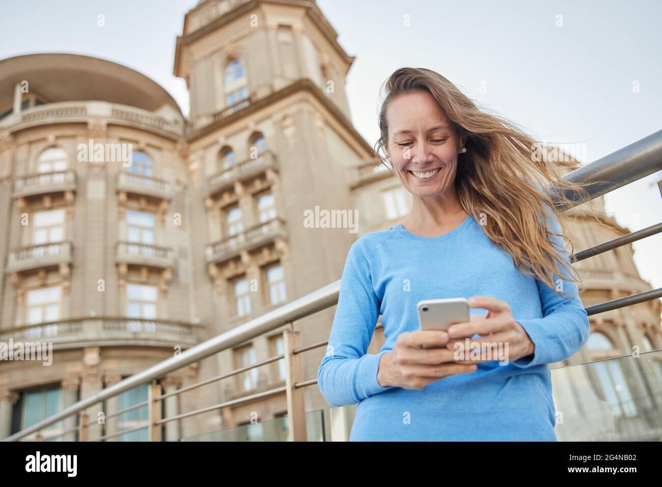 Da sotto di sorridente signora adulta in abiti casual in piedi vicino recinto e vecchio edificio, mentre navigando al telefono nel quartiere della città in luce del giorno Foto Stock