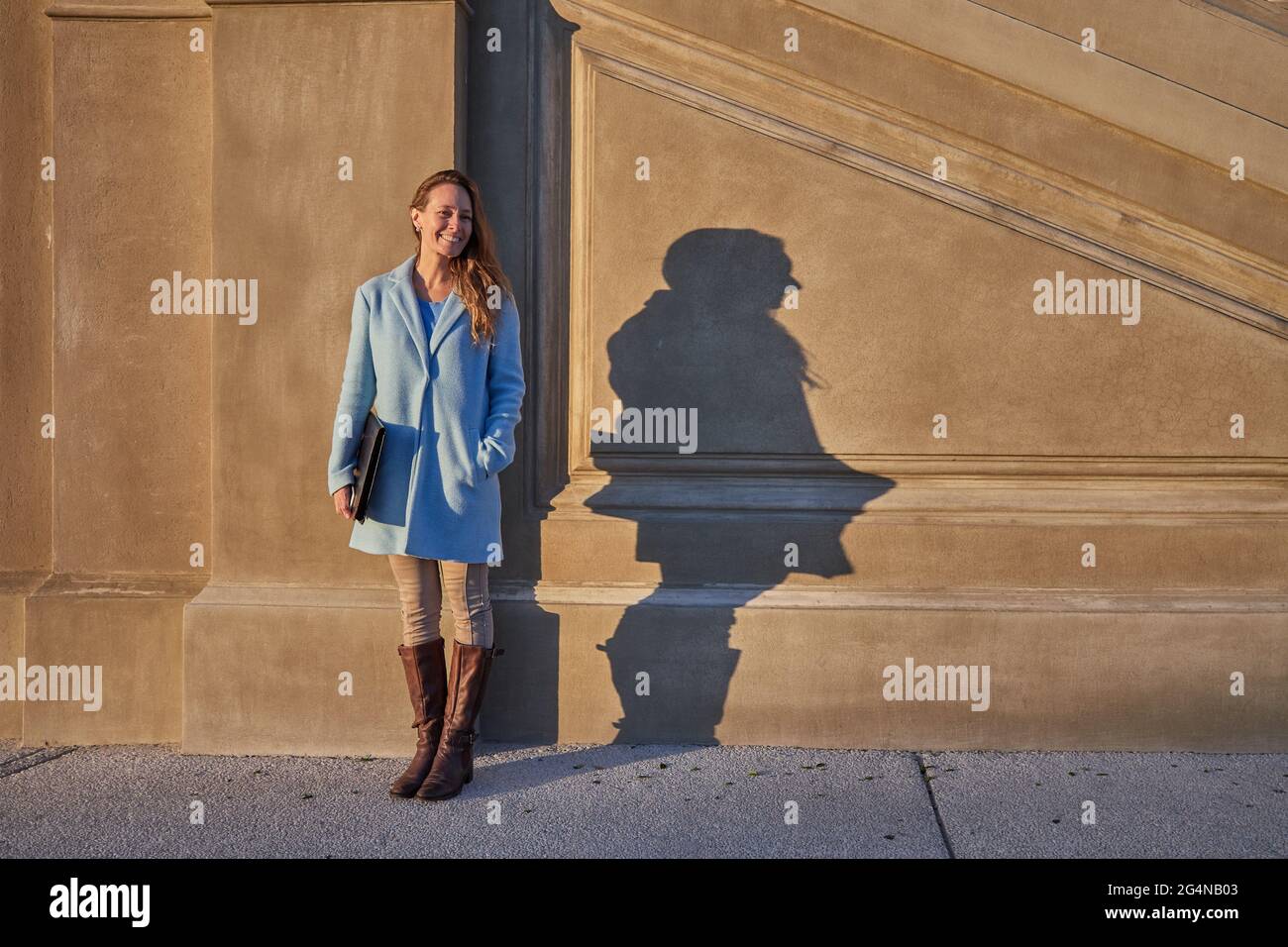 Corpo pieno di femmina felice adulta in cappotto blu in piedi vicino muro di costruzione di pietra con cartella in mani in città strada in giorno di sole Foto Stock