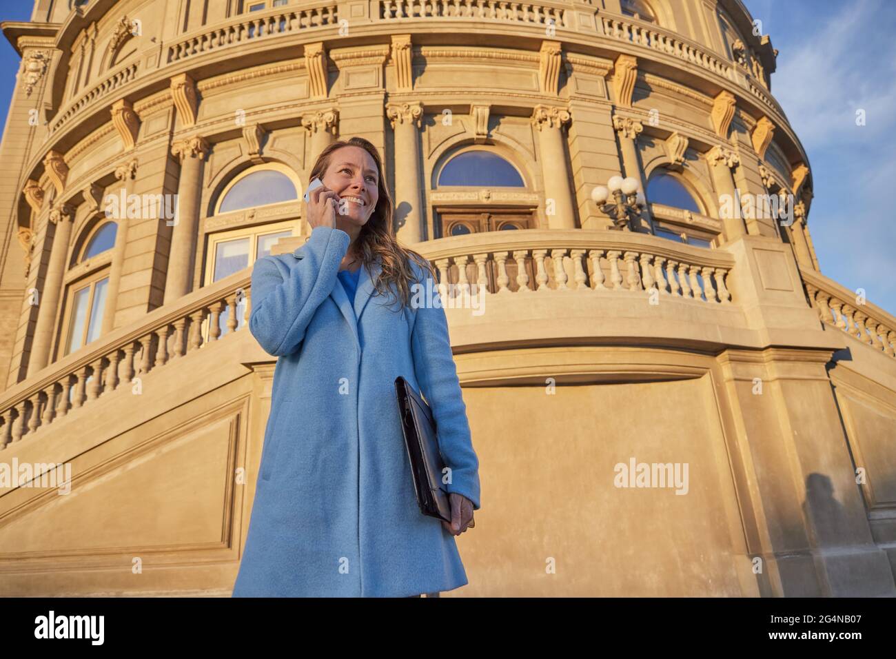 Felice adulto Donna in cappotto blu in piedi vicino a vecchio edificio mentre parlando cellulare nel quartiere della città in giorno di sole sotto cielo nuvoloso blu Foto Stock