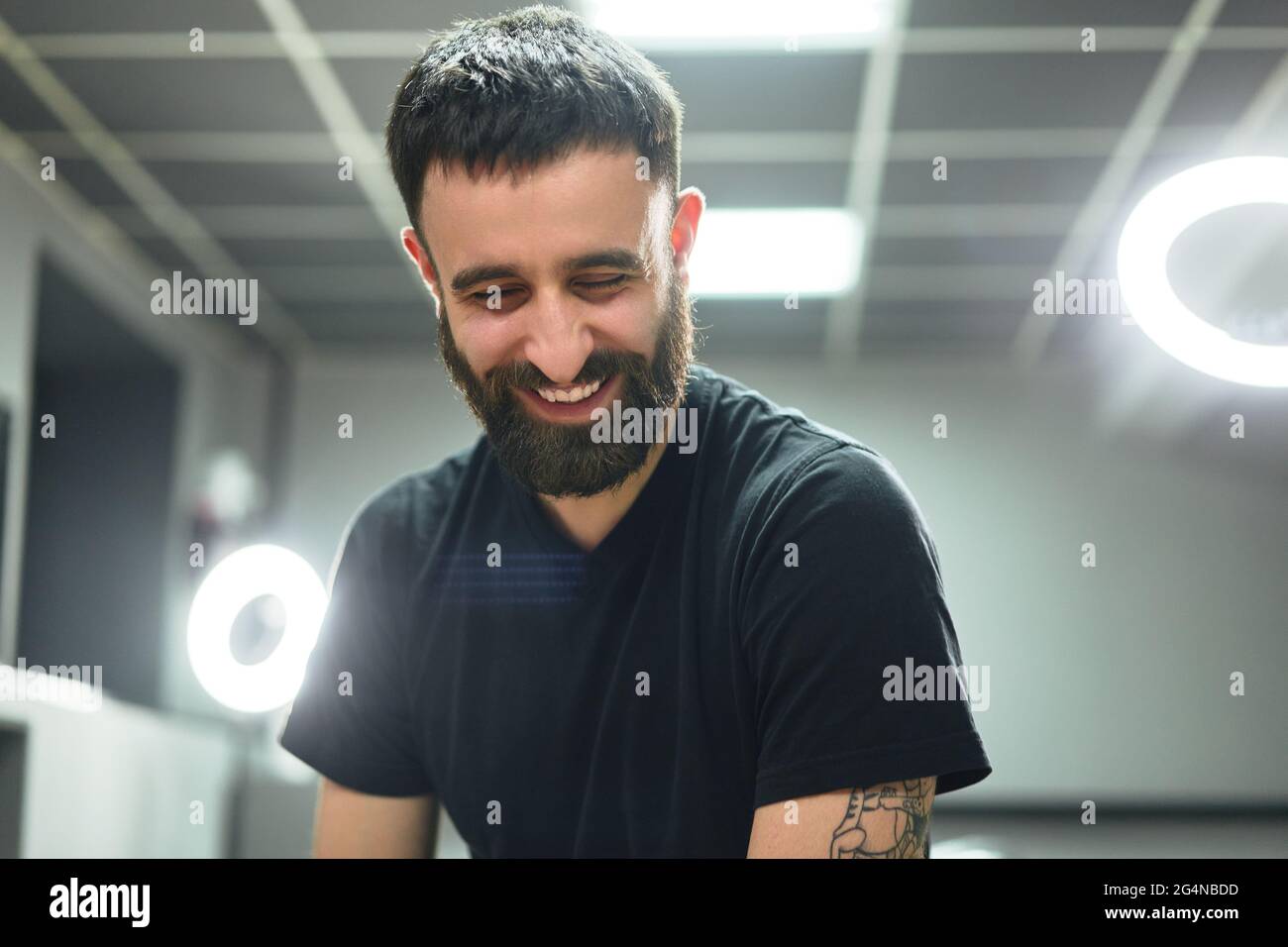 Tatuista maschile adulto con cuscinetti felici in abiti casual seduti su sgabello vicino a lampada ad anello e strumenti mentre si guarda verso il basso in un salone di tatuaggio leggero Foto Stock