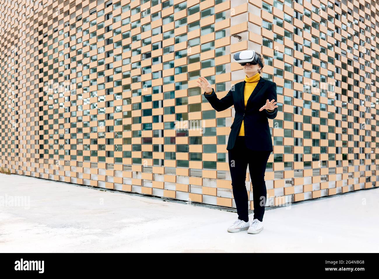 Femmina sorridente in giacca e dolcevita con occhiali VR vicino alle finestre dell'edificio in luce del giorno Foto Stock