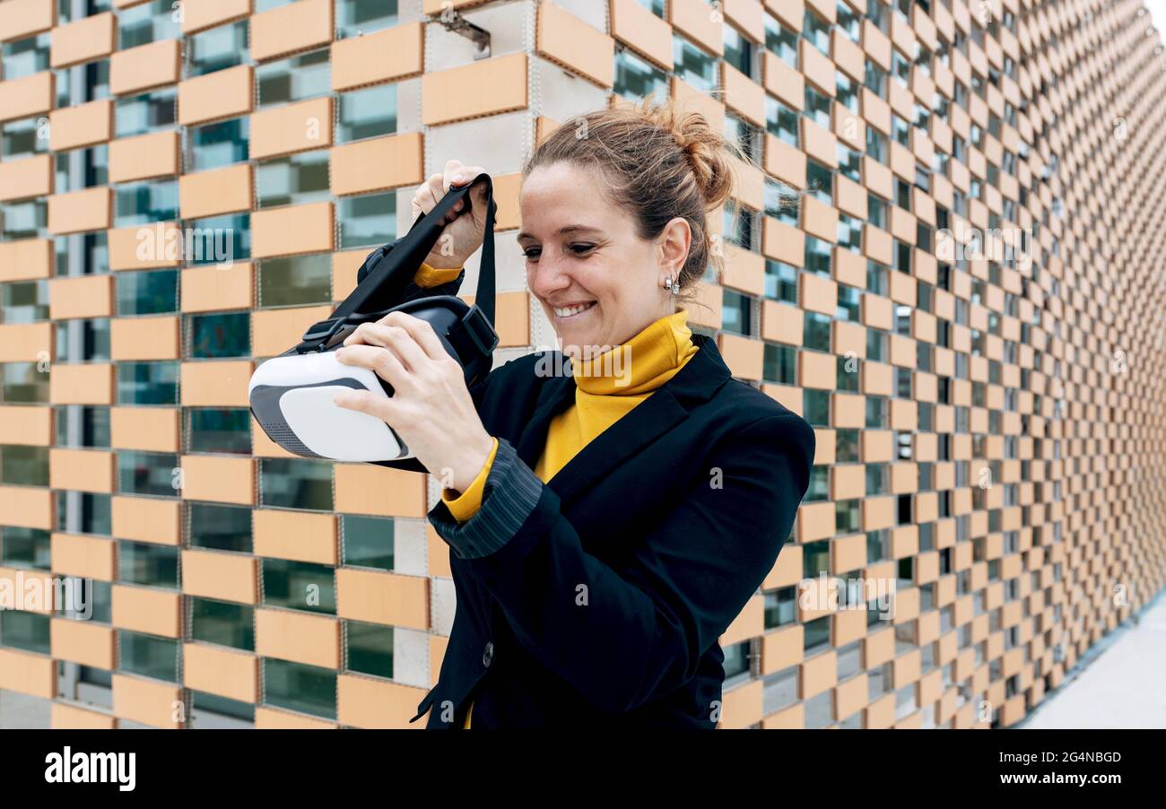 Donna adulta sorridente in un elegante abito che si trova vicino alla parete dell'edificio con occhiali VR e che guarda lontano dalla luce del giorno Foto Stock