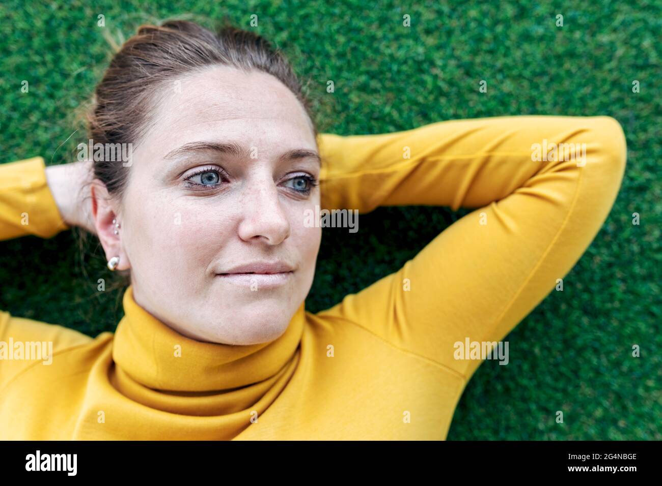 Dall'alto di sognante donna adulta in abiti casual giacente su erba verde con le mani dietro la testa e guardando via Foto Stock