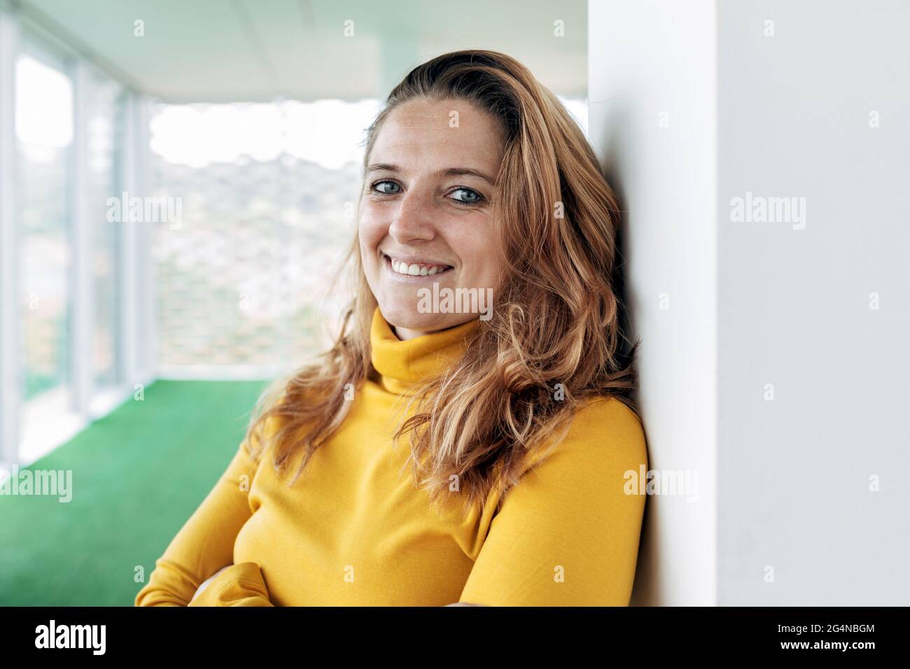 Donna adulta felice in blusa gialla guardando la macchina fotografica mentre si trova vicino colonna in un edificio luminoso con finestre panoramiche e pavimento verde Foto Stock