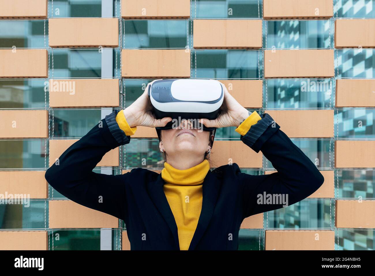 Donna in giacca e dolcevita con occhiali VR vicino alle finestre dell'edificio nella luce del giorno Foto Stock