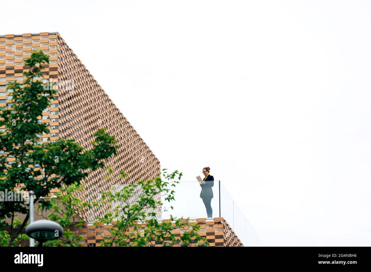 Da sotto di donna in abito elegante in piedi sul balcone di un edificio moderno con elementi geometrici sulle finestre mentre si usa un tablet vicino a ringhiera di vetro Foto Stock