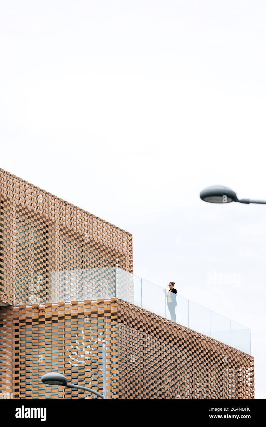Da sotto di donna in abito elegante in piedi sul balcone di un edificio moderno con elementi geometrici sulle finestre mentre si usa un tablet vicino a ringhiera di vetro Foto Stock