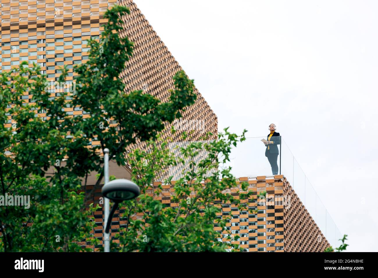 Da sotto di donna in abito elegante in piedi sul balcone di un edificio moderno con elementi geometrici sulle finestre mentre si usa un tablet vicino a ringhiera di vetro Foto Stock