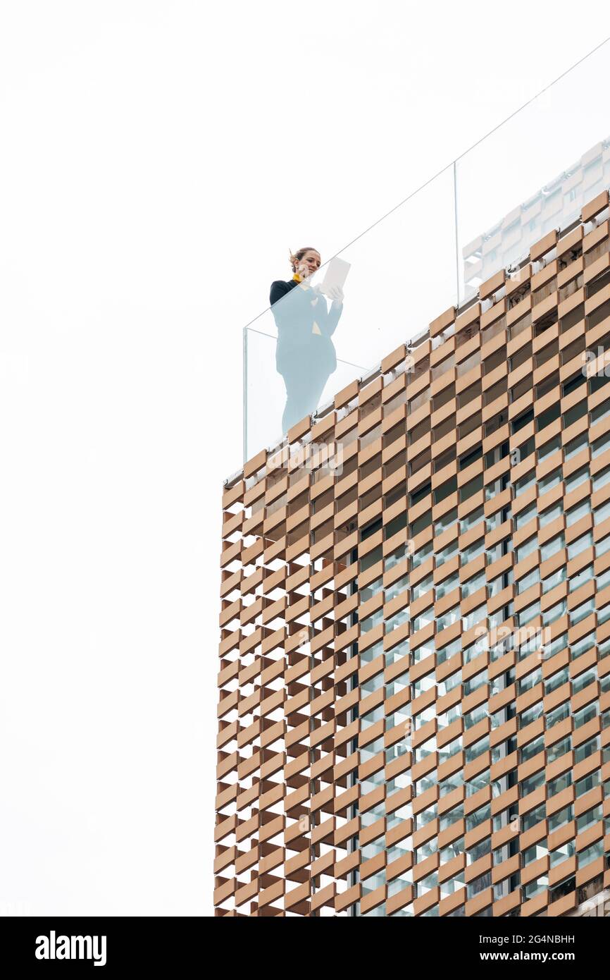 Da sotto di donna in abito elegante in piedi sul balcone di un edificio moderno con elementi geometrici sulle finestre mentre si usa un tablet vicino a ringhiera di vetro Foto Stock