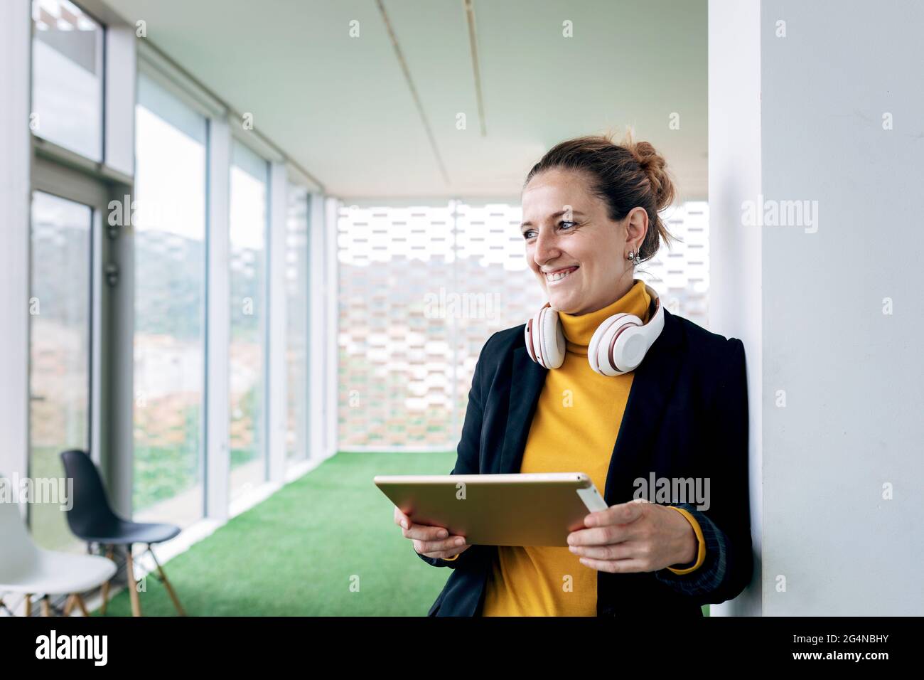 Donna adulta sorridente in un abito elegante con cuffie e tablet appoggiati sulla colonna in un edificio luminoso vicino alle finestre mentre si guarda via Foto Stock