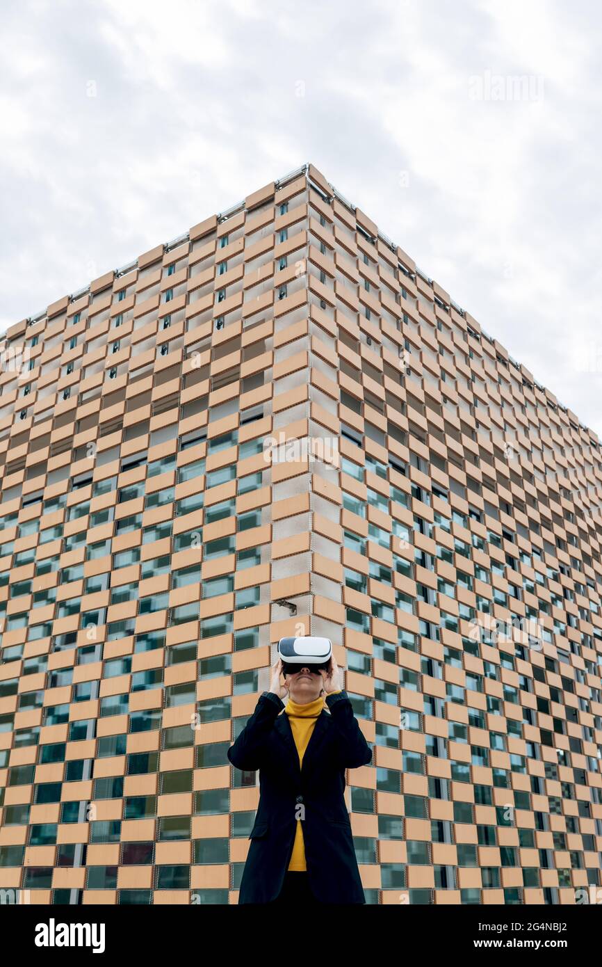 Donna in giacca e dolcevita con occhiali VR vicino alle finestre dell'edificio nella luce del giorno Foto Stock