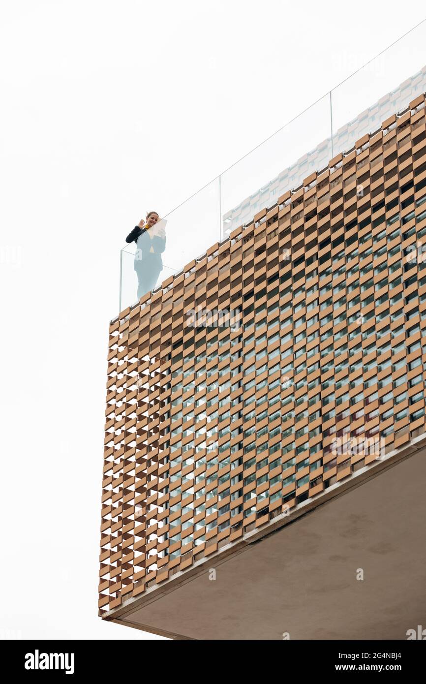 Da sotto di donna in abito elegante in piedi sul balcone di un edificio moderno con elementi geometrici sulle finestre mentre si usa un tablet vicino a ringhiera di vetro Foto Stock