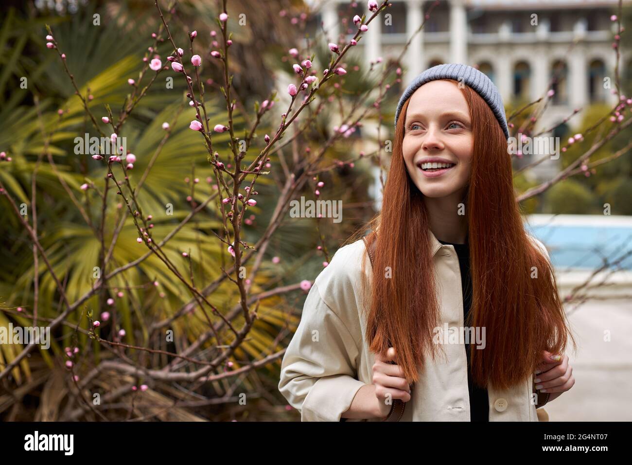 Sorprendente giovane donna dai capelli rossi in cappello che si pone in fiore albero a primavera. Bella giovane donna felice godendo l'odore nel giardino fiorente della molla, da sola, Foto Stock