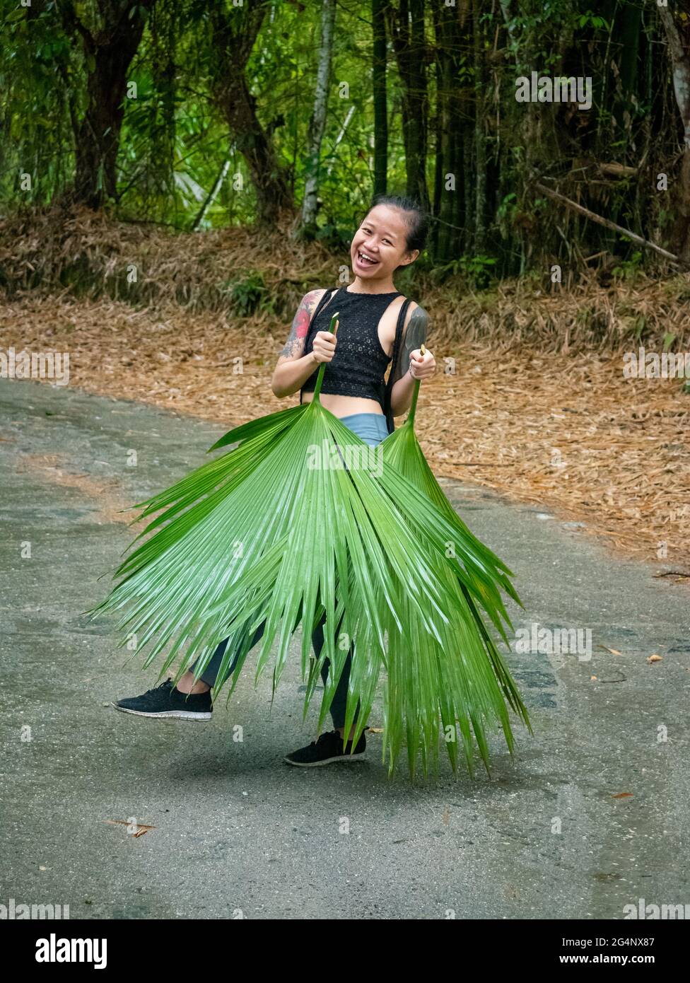 Piccola donna asiatica che copre le gambe con foglie enormi, ballando e guardando la macchina fotografica a Minca, Colombia Foto Stock