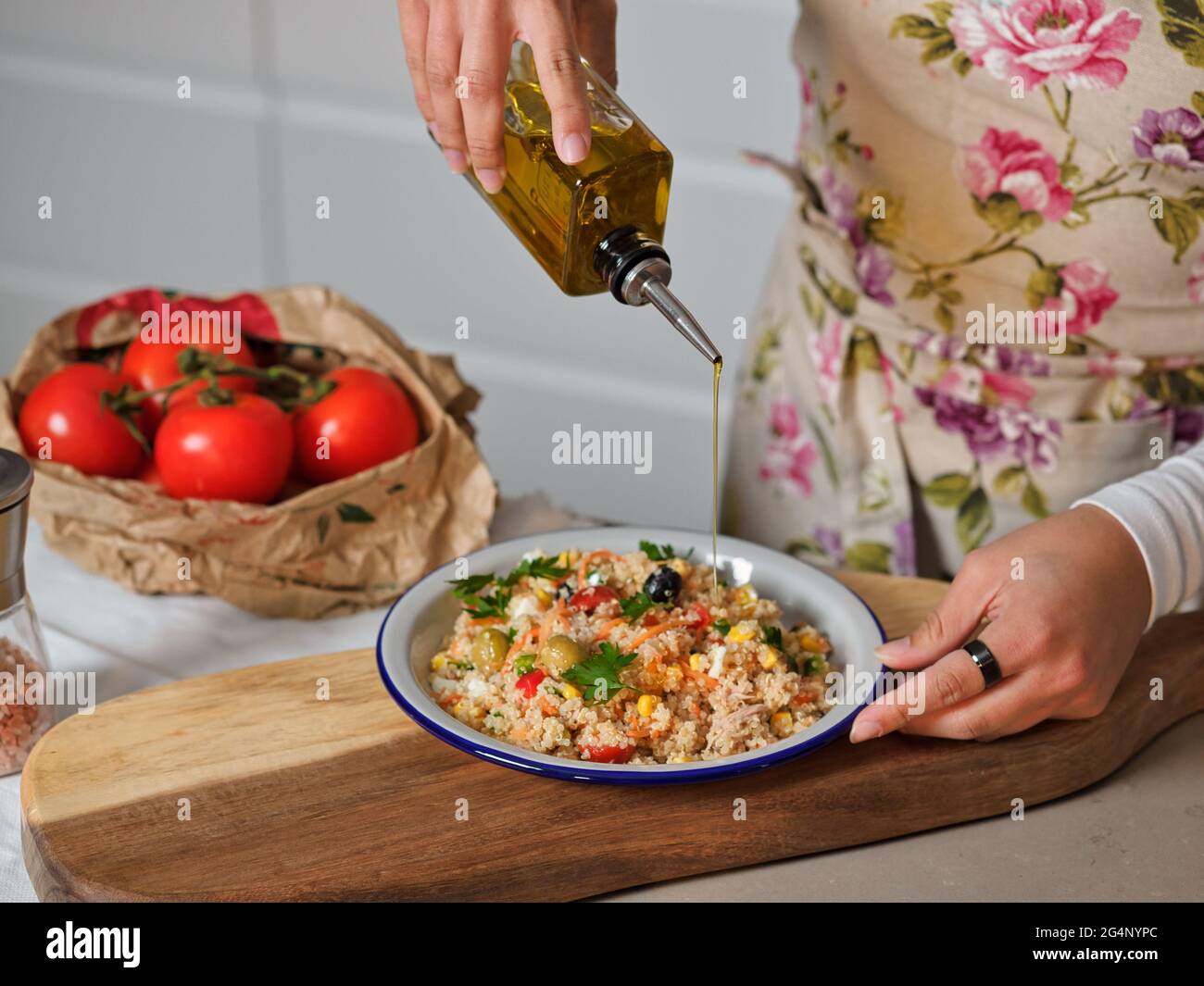 le mani della donna leccano l'olio d'oliva sul piatto di insalata di quinoa in cucina domestica, fondo di sacchetto di carta con pomodori e cassetti bianchi della cucina Foto Stock