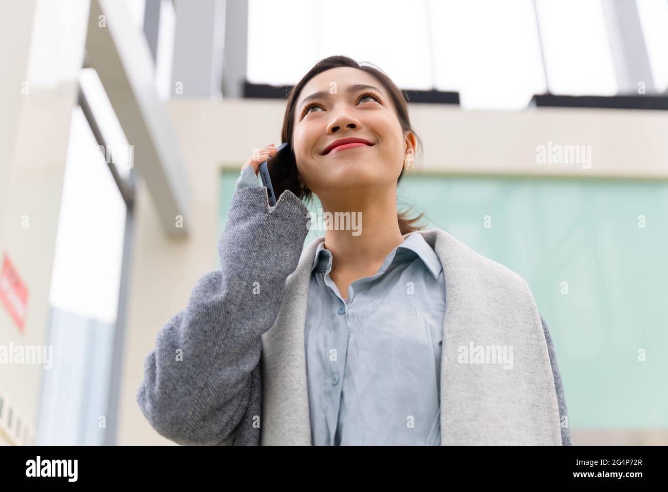 Foto a basso angolo di giovane bella donna d'affari asiatica che chiama con il telefono cellulare in ufficio edificio con spazio di copia Foto Stock