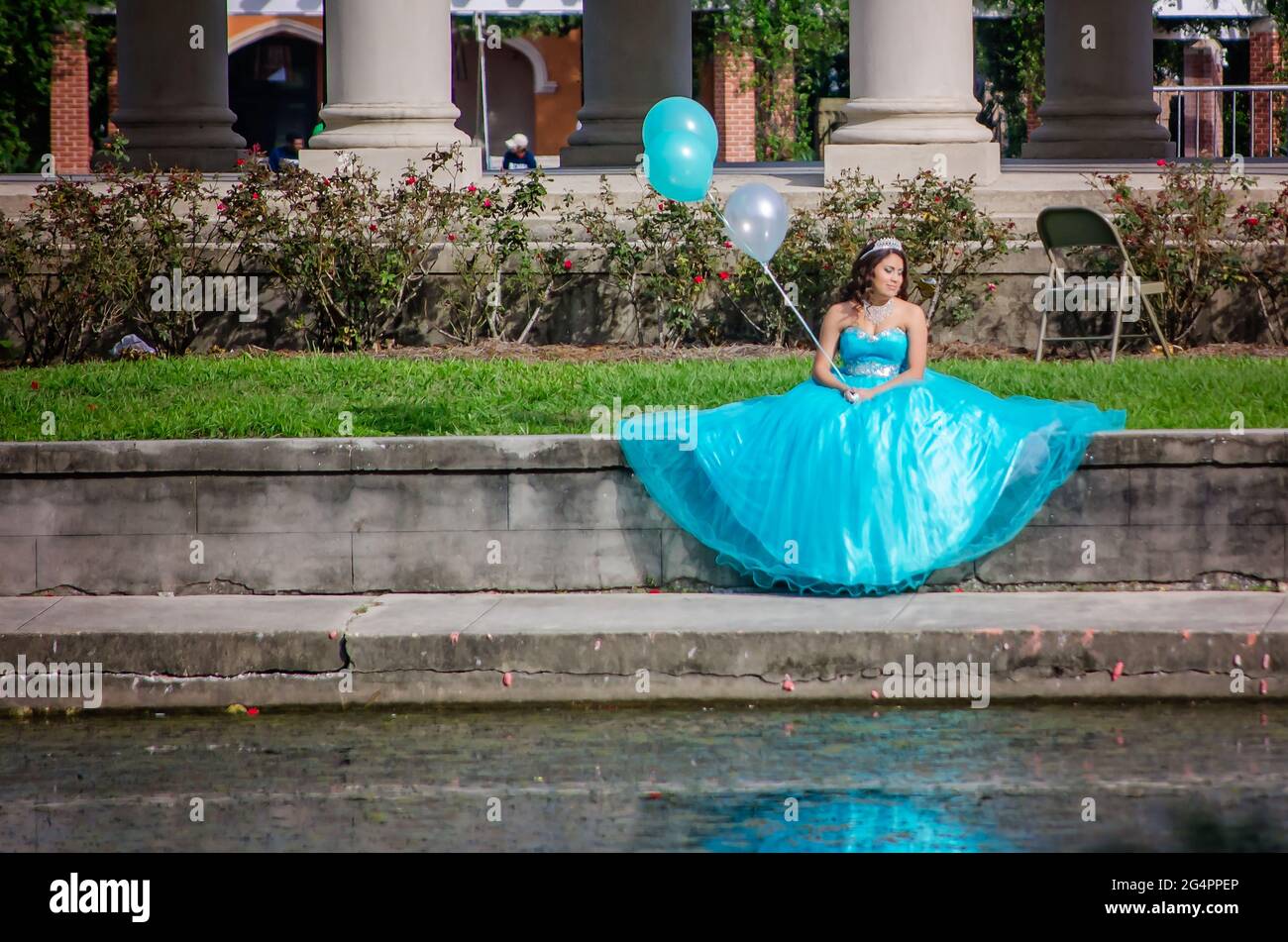 Una ragazza che tiene i palloncini mentre indossa un abito da palla e tiara siede sul lato dell'acqua al Peristyle nel New Orleans City Park a New Orleans, Louisiana. Foto Stock