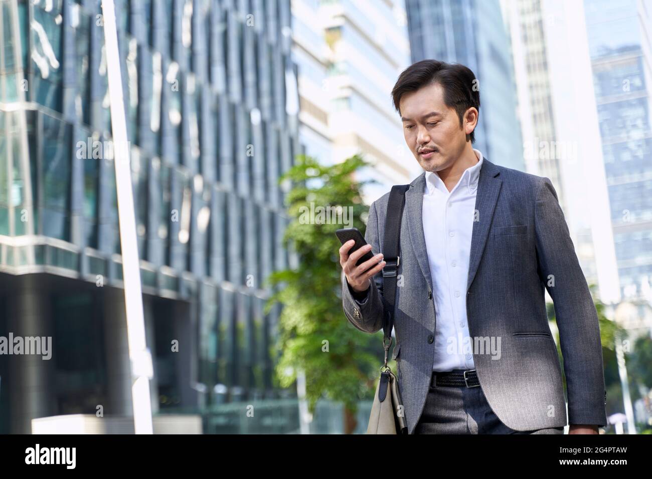 uomo d'affari asiatico che guarda il telefono cellulare mentre si cammina per lavorare nel quartiere finanziario della città moderna Foto Stock