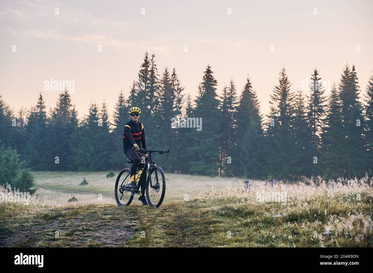 Ciclista maschile in bicicletta tuta ciclistica in mountain bike con alberi di conifere sullo sfondo. Uomo in bicicletta indossando casco e occhiali di sicurezza mentre si fa un giro in bicicletta in montagna al mattino. Foto Stock