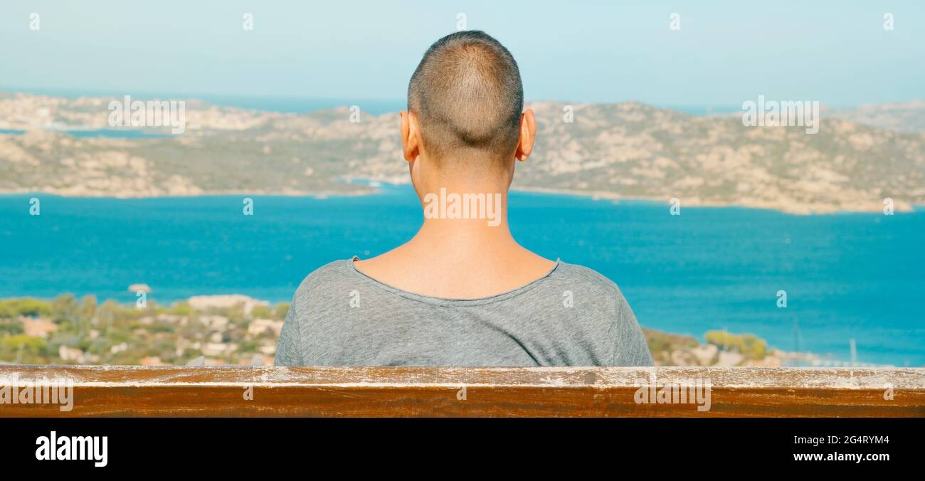 Primo piano di un giovane seduto in una panchina a Palau, sulla costa settentrionale della Sardegna, Italia, osserva il Mar Mediterraneo e la Maddalena Archipel Foto Stock