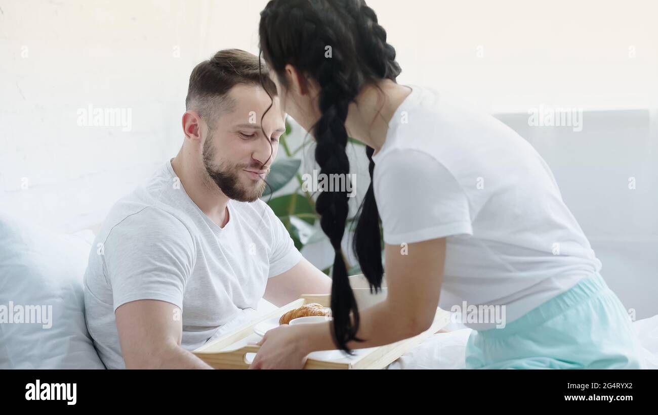 donna di brunette con trecce che porta il vassoio della colazione al ragazzo a letto Foto Stock