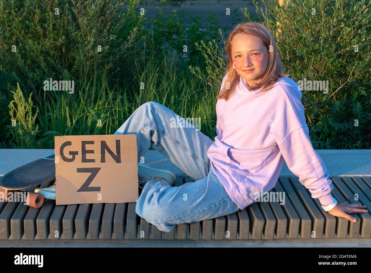 Una ragazza adolescente elegante in un jeans porpora con cappuccio e capelli rosa si siede su una panchina accanto a una tavola lunga e un poster con l'iscrizione Generation Z. GEN Foto Stock