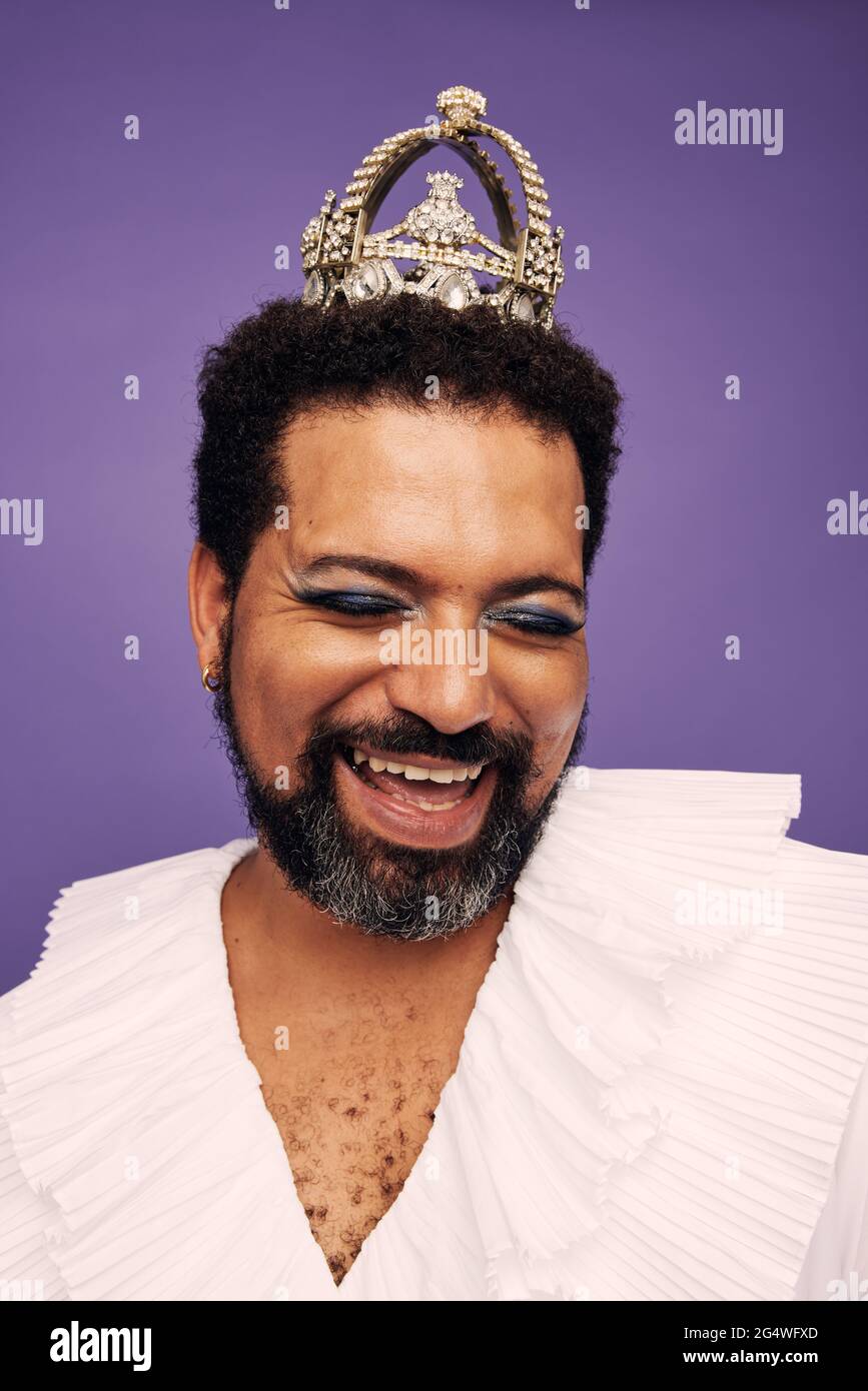 Uomo con barba che indossa trucco e corona. Ritratto di una regina di trascinamento felice su sfondo viola. Foto Stock
