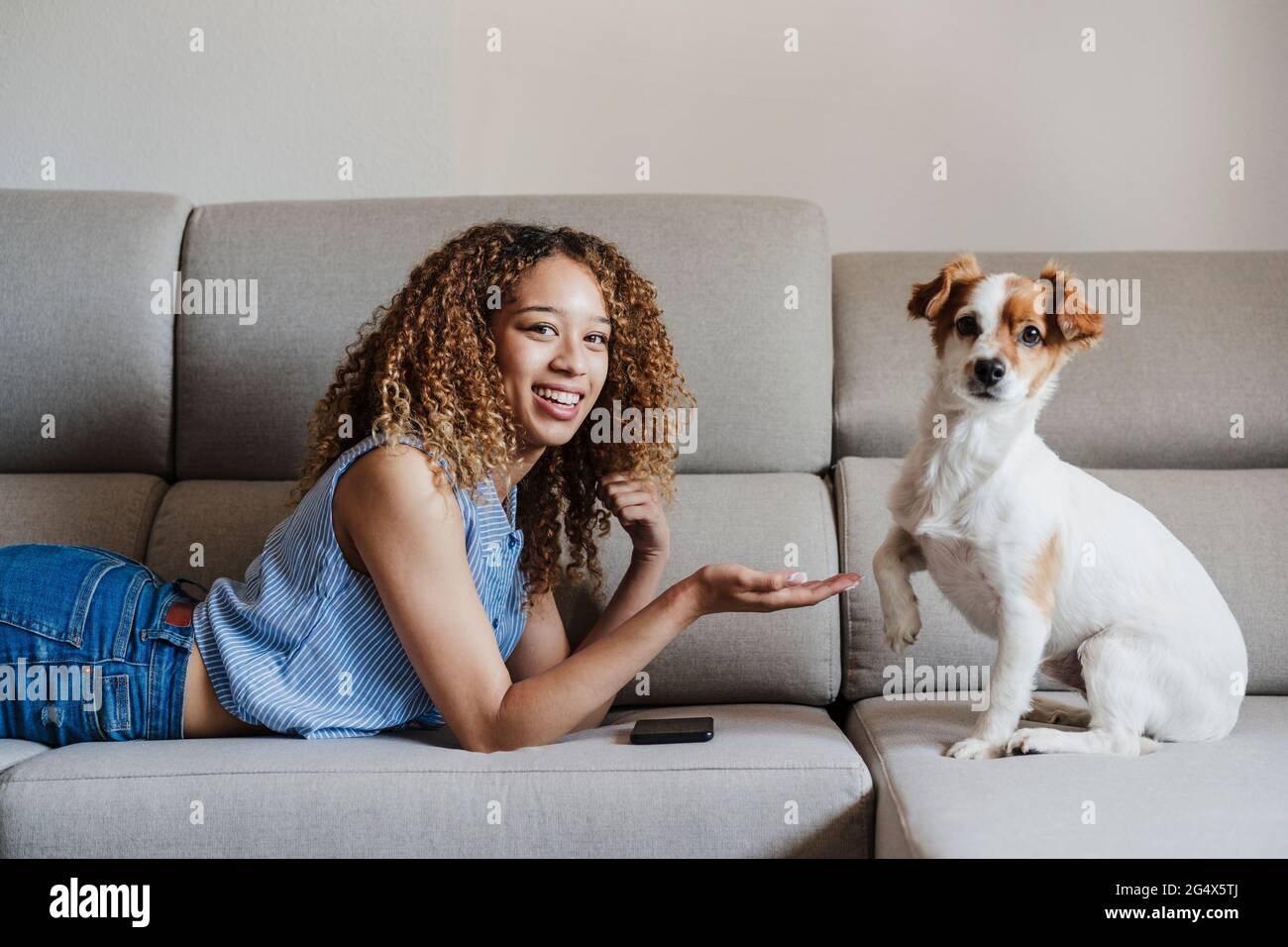 Giovane donna sorridente che offre la stretta di mano al cane sul divano a casa Foto Stock