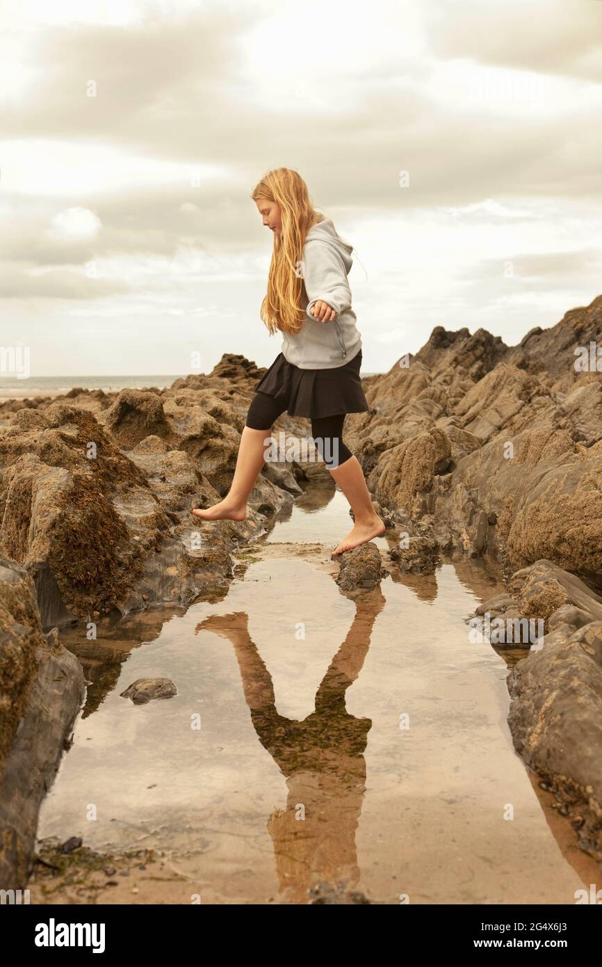 Ragazza bionda che salta sulla formazione rocciosa sopra la piscina di marea alla spiaggia Foto Stock