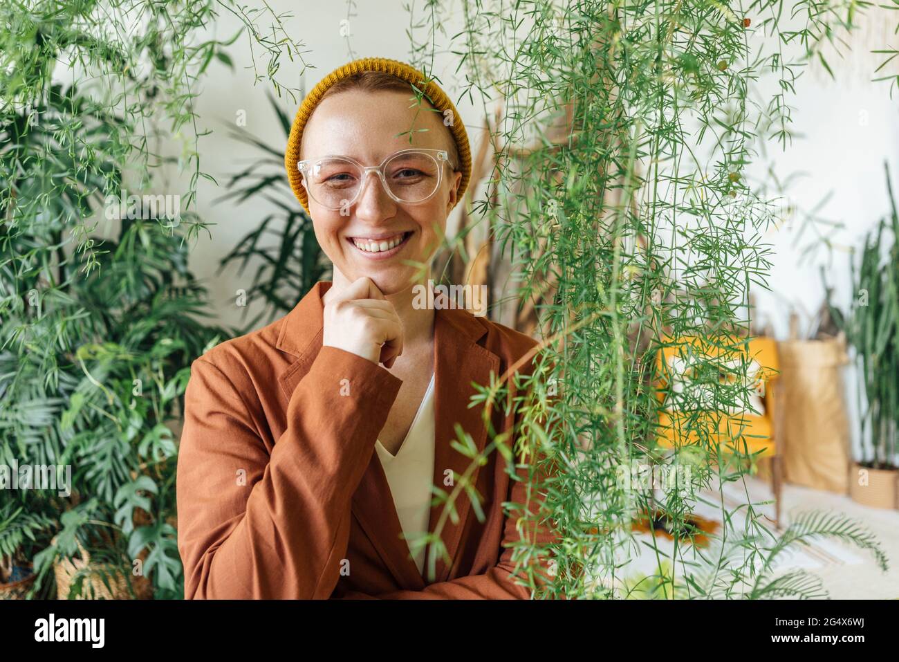 Imprenditore femminile sorridente con mano sul mento in piedi da piante sul posto di lavoro Foto Stock