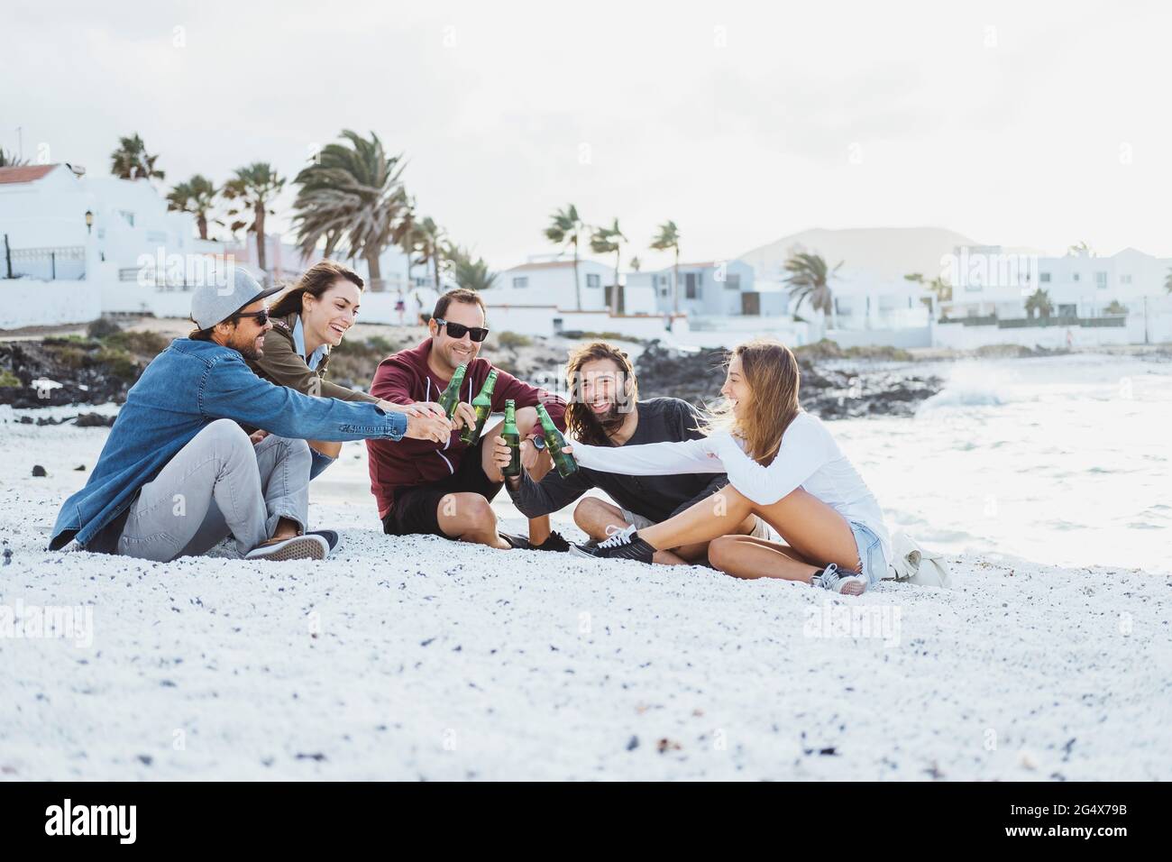 Amici maschili e femminili che tostano le bottiglie di birra mentre si siedono sulla spiaggia Foto Stock