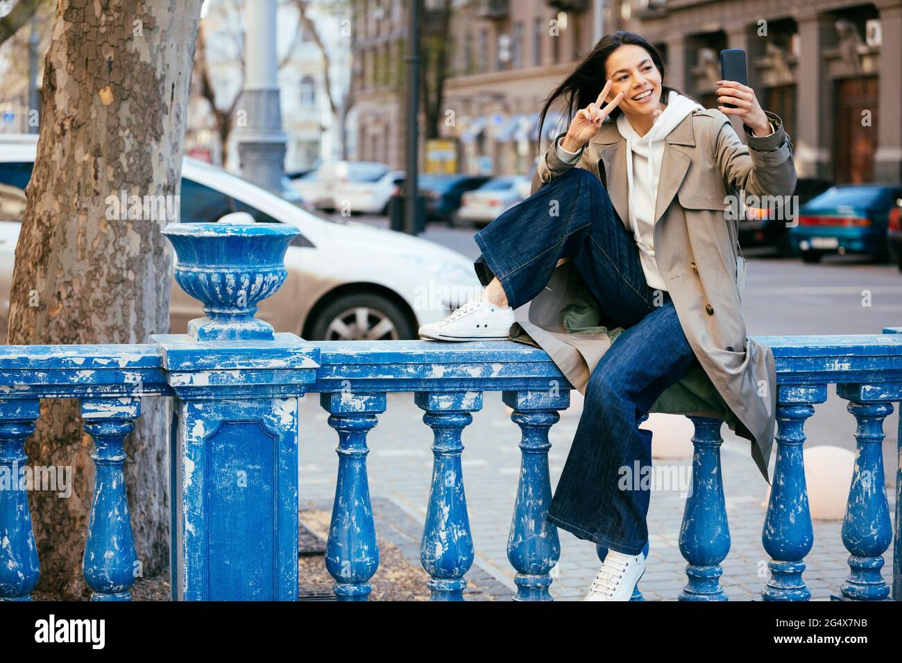 Donna sorridente seduta sulla ringhiera blu che mostra il segno di pace mentre prende selfie Foto Stock