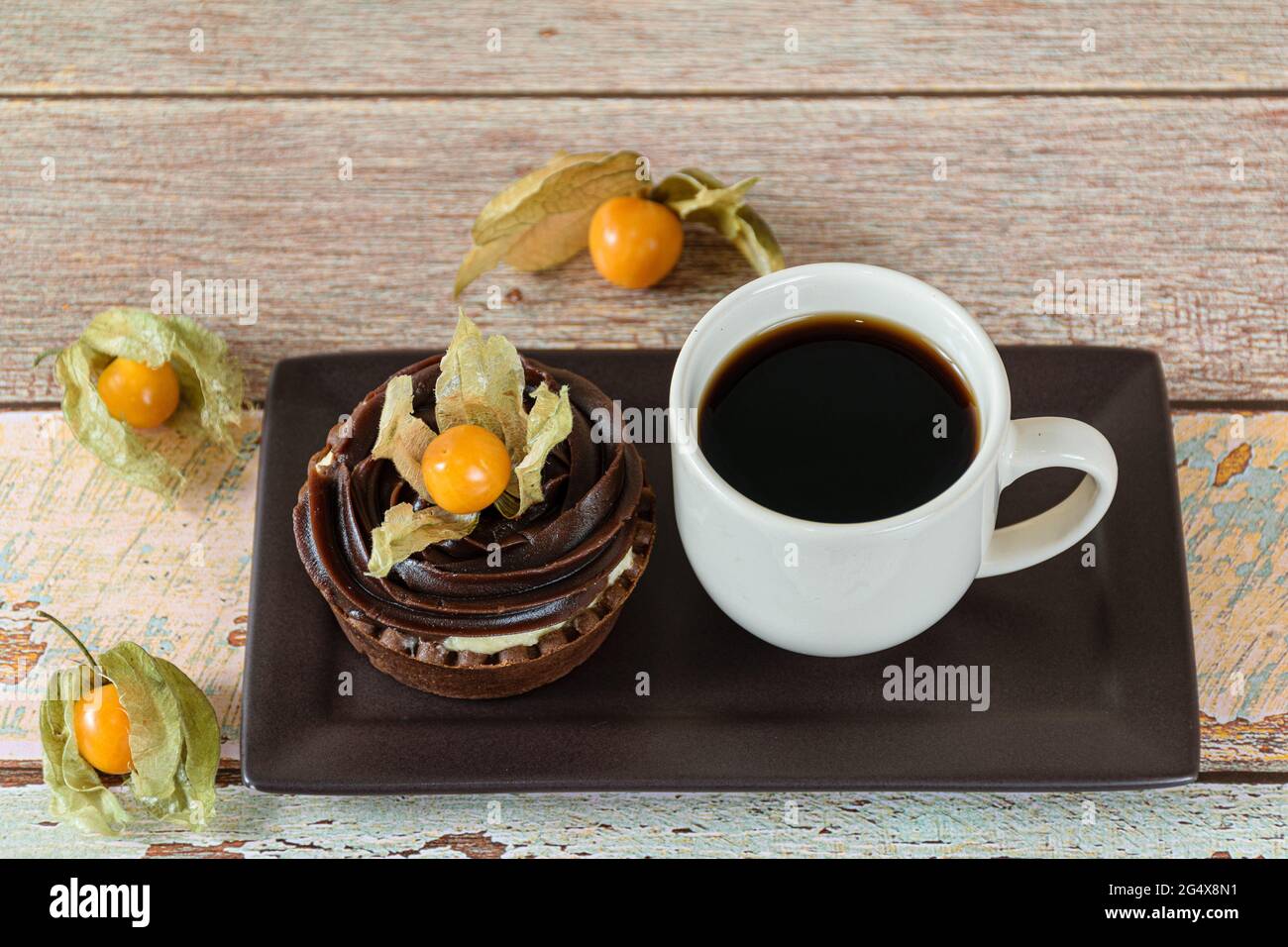 Mini torta con crema di mascarpone e ganache al cioccolato, decorata con physalis, accanto ad una tazza di caffè. Foto Stock