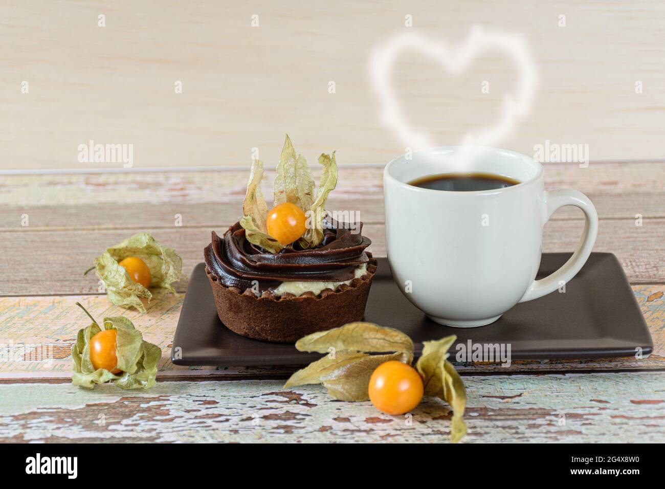 Mini torta con crema di mascarpone e ganache al cioccolato, decorata con physalis, accanto ad una tazza di caffè (con cuore di fumo). Foto Stock