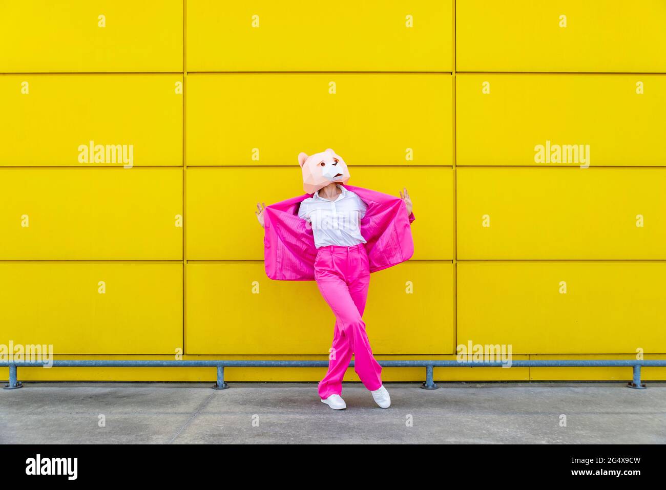 Donna che indossa un abito rosa vivace e una maschera dell'orso in piedi davanti alla parete gialla con giacca a stendimento Foto Stock