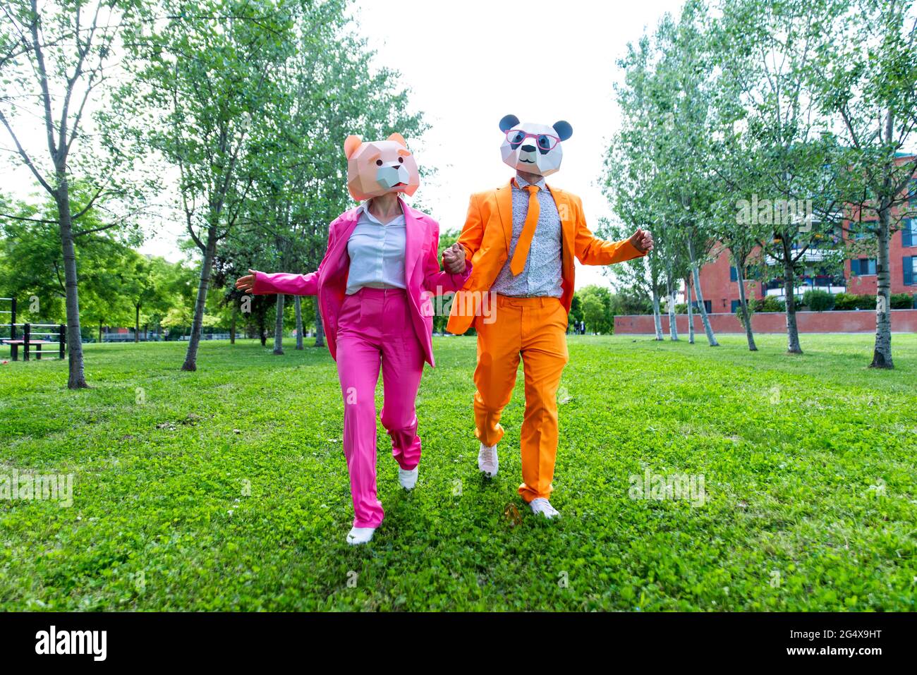 coupleÂ adulto indossa abiti vivaci e maschere animali che tengono le mani mentre corrono insieme attraverso il parco verde Foto Stock