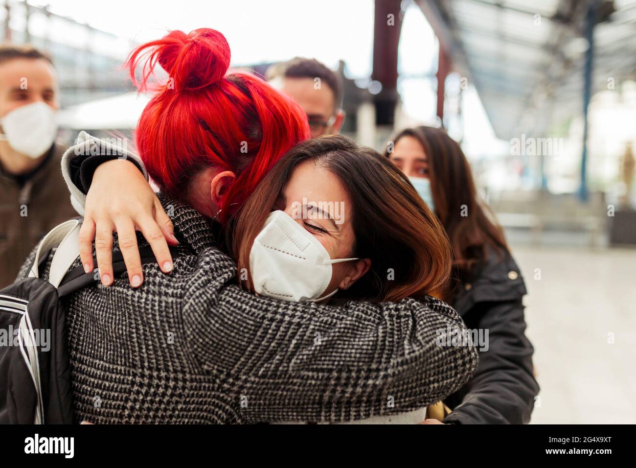 Le amiche in maschere di faccia abbracciano alla stazione ferroviaria durante COVID-19 Foto Stock