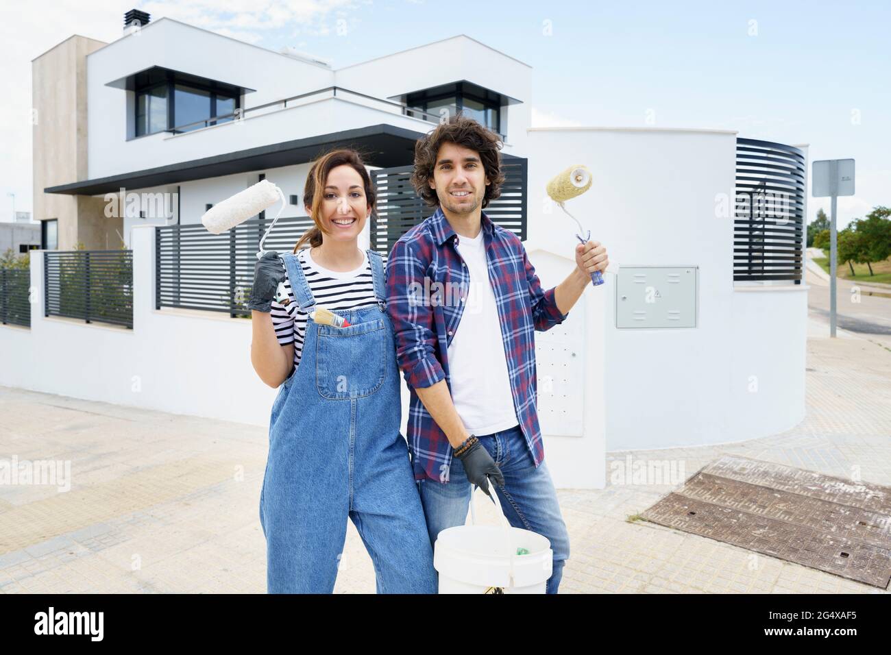 Coppia sorridente con rullo di vernice in piedi di fronte a casa nuova Foto Stock